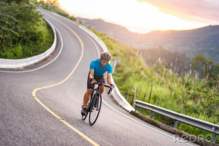 Canvas asian male riding on a black bicycle along the winding road up a hill, wearing a cycling blue jersey, crash helmet and goggles, sunset light, grey sky, and forest trees and mountains in the background