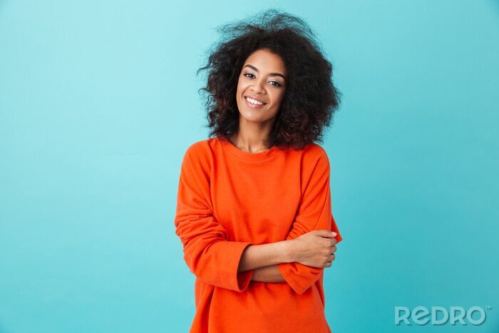 Canvas Colorful portrait of amazing woman in red shirt with afro hairstyle looking on camera with smile, isolated over blue background