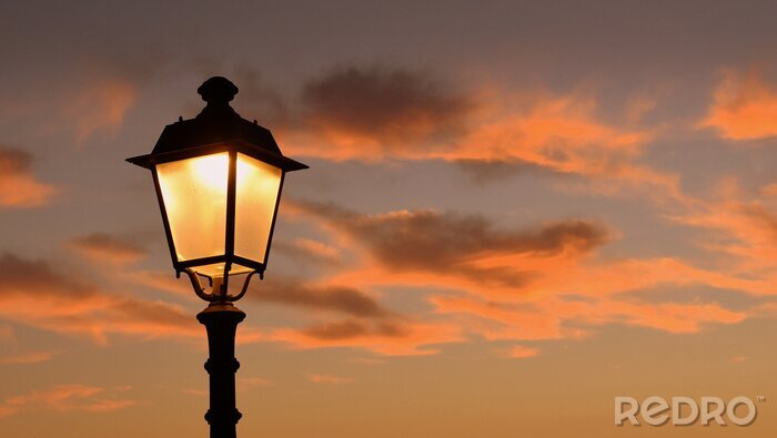 Canvas Oranje gloeiende avondlucht, omlijst door takken en antieke lantaarn op de achtergrond
