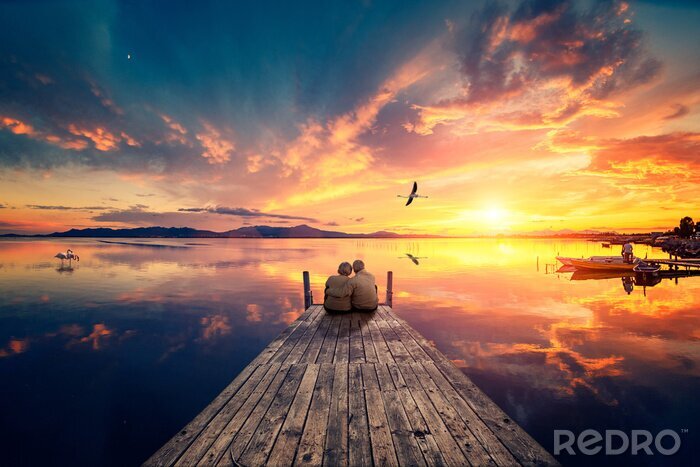 Canvas Senior couple seated on a wooden jetty, looking a colorful sunset on the sea with a flying flamingo reflected on the calm water.