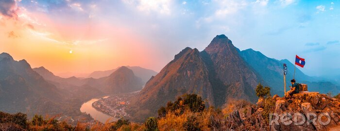 Canvas Sunset panoramic view of couple of trekkers sitting on a rock on top of Nong Khiaw View Point with beautiful mountain and Nam Ou river in background, Luang Prabang Province, Northern Laos.