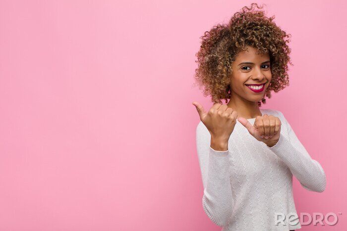 Canvas young african american woman smiling cheerfully and casually pointing to copy space on the side, feeling happy and satisfied against pink wall