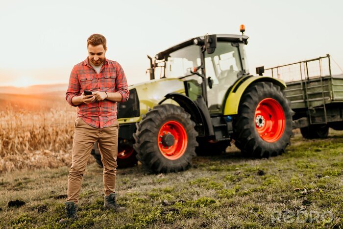 Fotobehang Mannelijke landbouwer die aan gebied werken die smartphone in moderne landbouw gebruiken - tractorachtergrond