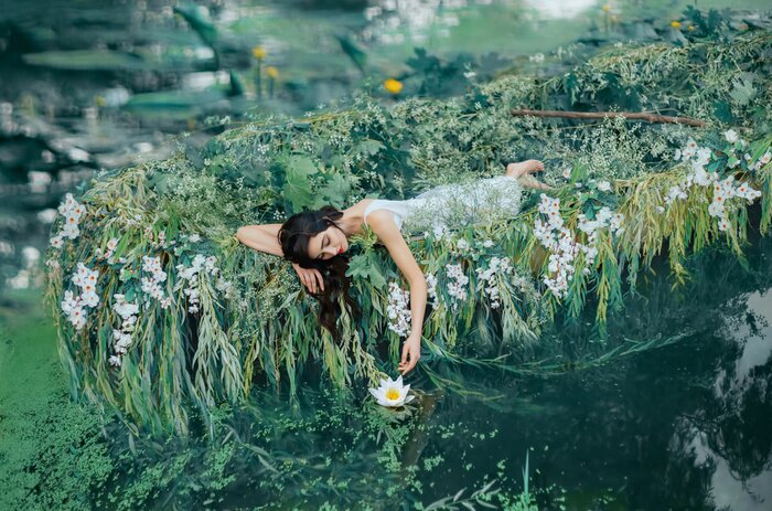 Poster A brunette woman with long hair lies in a boat and gently touches a water lily her hand. The princess in a white dress floats on the lake. Friday, relaxation and enjoyment. Creative wedding decor
