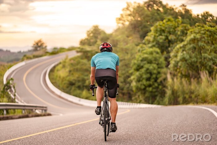 Poster back view of a cyclist on top of a mountains winding road, riding a black bicycle down a hill, wearing bike helmet and blue cycling jersey, with grey clouds sunset sky and forest in the background.