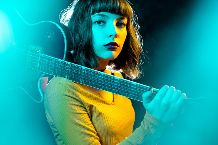 Poster Beautiful young hipster woman with curly hair with red guitar in neon lights. Rock musician is playing electrical guitar. 90s style concept.