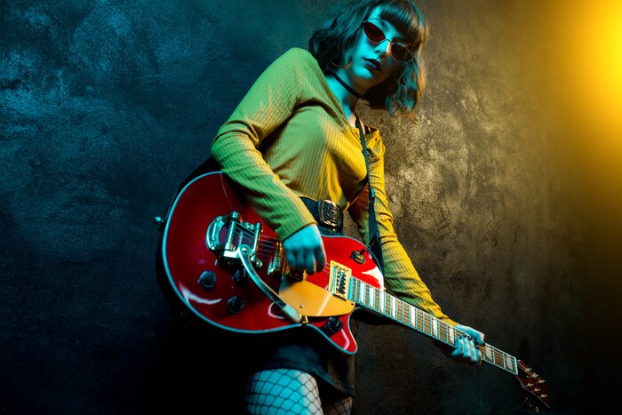 Poster Charming hipster woman with curly hair with red guitar in neon lights. Rock musician is playing electrical guitar. 90s style concept.