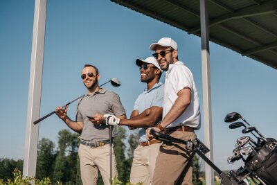 Drie lachende mannen in zonnebril houden golfclubs buitenshuis