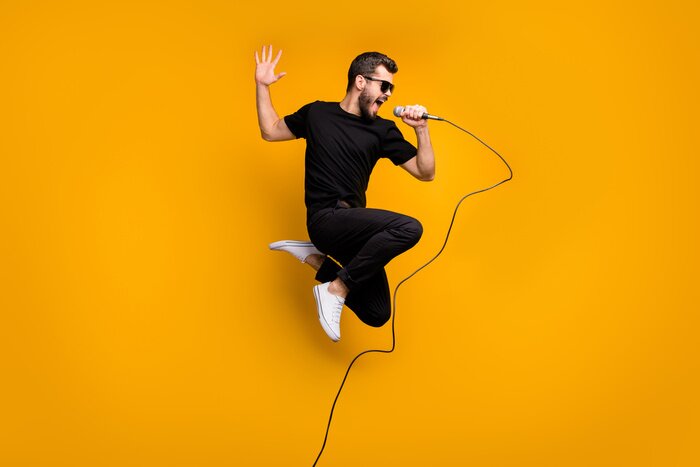 Poster Full body profile photo of crazy hipster guy jumping high holding microphone music lover singing favorite song wear sun specs black t-shirt pants isolated yellow color background