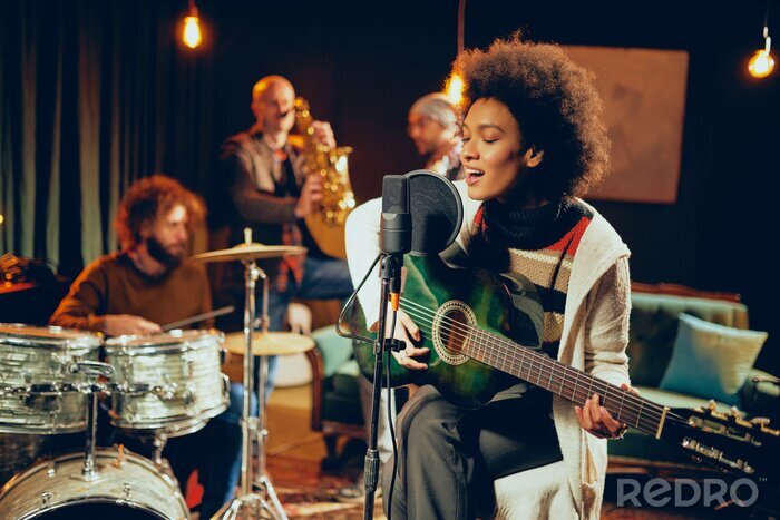 Poster Mixed race woman singing and playing guitar while sitting on chair with legs crossed. In background drummer, saxophonist and bass guitarist.