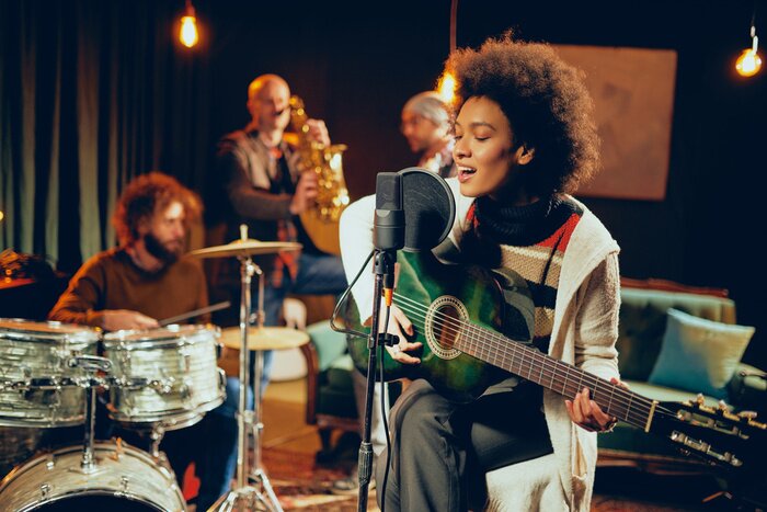 Poster Mixed race woman singing and playing guitar while sitting on chair with legs crossed. In background drummer, saxophonist and bass guitarist.