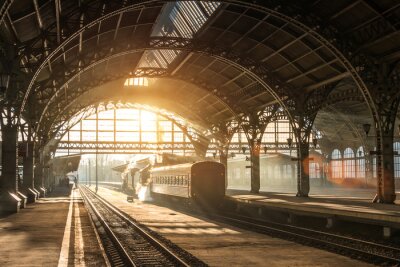 Poster Old railway station with a train and a locomotive on the platform awaiting departure. Evening sunshine rays in smoke arches.
