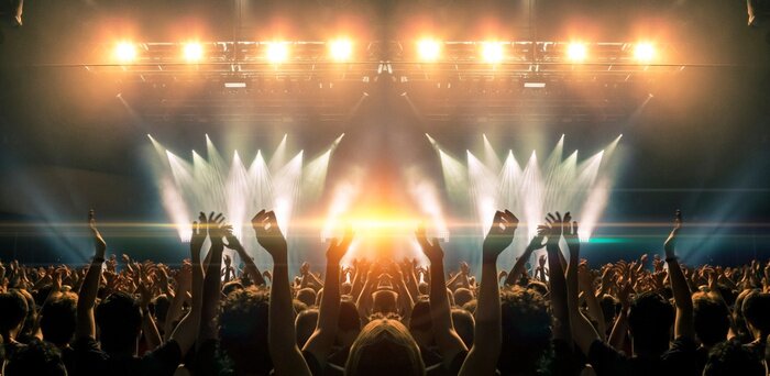 Poster Photo of a concert hall with people silhouettes clapping in front of a big stage lit by spotlights. Shot is taken from concert crowd point of view, lens flare is visible.