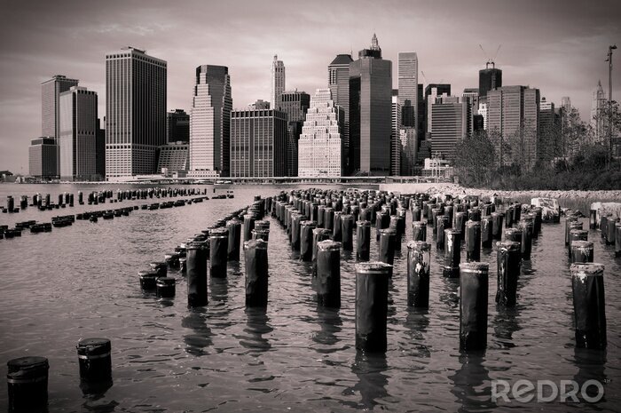 Poster Skyline von Manhattan mit Holzpfählen im Wasser, Schwarzweiß